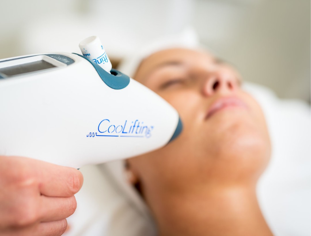 CoolLifting Behandlung: Eine Kundin liegt mit geschlossenen Augen und weißem Haarband, während die Behandlerin ein Beauty-Gun-Handstück an der Wange führt